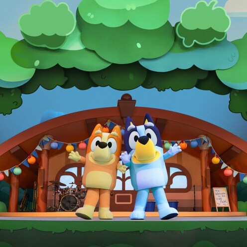 Bingo and Bluey pose on a stage set designed to look like a house in the woods at Bluey’s Best Day Ever
