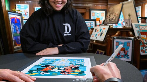 An artist signing a print featuring Disney characters for a Guest in an art gallery