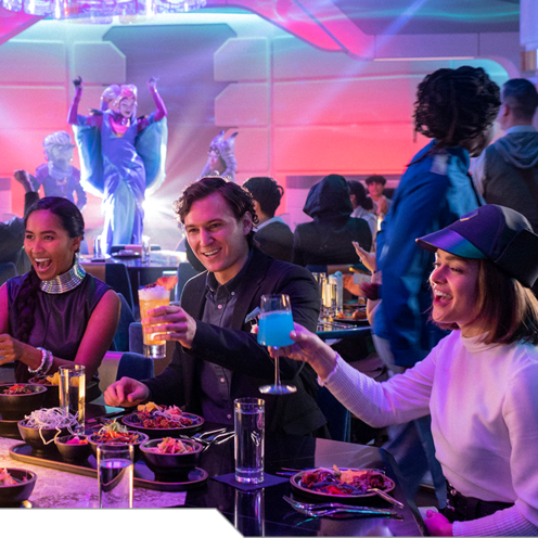 Guests raise a toast at a table while an intergalactic entertainer performs on stage in the background aboard the Halcyon starcruiser