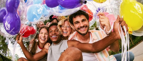 Un groupe de cinq amis s’assoit étroitement ensemble, souriant joyeusement, entouré de ballons en forme de Mickey Mouse