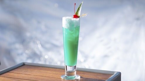 A tall tapered glass containing a Blue Hawaiian cocktail on the rocks is garnished with a maraschino cherry and a pointed leaf skewered by a miniature plastic sword
