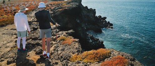 Two people wearing hats hike along a rocky clifftop high above the sea