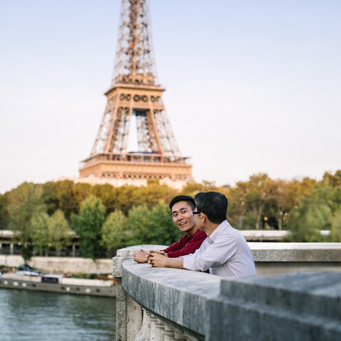 Two men stand with their arms resting on a cement railing overlooking the Seine River with the Eiffel Tower in the background
