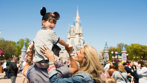 A boy wearing Mickey ears sits on top of his father’s shoulders in front of Cinderella Castle