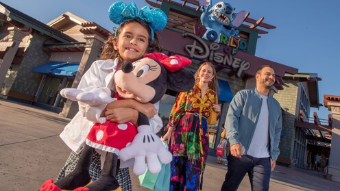 A girl hugging a large Minnie Mouse plush walking with her parents and a large Stitch figure above a sign with the words ‘World of Disney