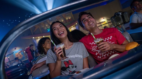 Guests smiling and holding beverages while sitting in a car at the Sci Fi Dine In Theater Restaurant