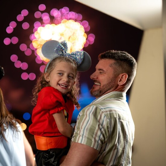 A father holds his daughter in his arms while they smile as fireworks burst behind them