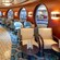 A lounge has high stools at a casual bar and rows of chairs with cocktail tables next to porthole windows