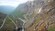 A paved road zig zags down a hillside, over a waterfall and into a valley with steep walls