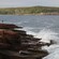 A person stands on rocks above the splashing surf near a rocky coastline covered in trees