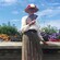 A woman dressed in Victorian attire holding a fan next to a flowerbed
