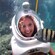 A helmet diver smiles as a fish swims in front of her underwater