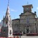An old stone building with a clock tower sits across the street from a cathedral shaped stone fountain