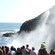 A crowd watches a large spray of water against a rocky cliff