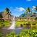 Water sprays from the middle of a lagoon, surrounded by palm trees, tropical plants and grass lawn