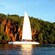 A luxury catamaran glides through the water alongside a verdant cliff