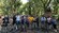 Un grupo de visitantes con bicicletas sonríen a la cámara en una pasarela pavimentada y altamente poblada en un parque grande