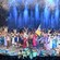 A group of performers dressed in 19th century attire on a stage with fireworks and fountains of water