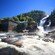 A waterfall falls between 2 hills and spills into a pool of raging water near a building