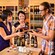 A man pours wine into a tasting cup for a woman who stands near her friend and shelves of wine bottles