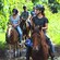 People wearing helmets and riding saddled horses through an area with lush greenery