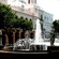 A fountain with 4 statues placed evenly around the rim sits in a plaza lined by trees