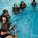 A group of scuba students stand in a swimming pool and gather around a dive instructor while learning how to use their equipment