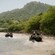 Off road buggies driving through a shallow river with a forested mountain in the background