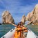 A couple wearing life vests smiling with arms raised while on a boat near El Arco of Cabo San Lucas rock formations