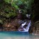A cascading waterfall emptying into a turquoise natural pool, surrounded by jungle cliffs and a wooden staircase