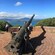A cannon mounted on a circular brick platform overlooking the ocean with lush greenery and distant mountains