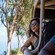 A smiling woman leaning out of a safari style vehicle on a road with trees and the ocean in the background