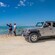 Two people taking a selfie on a beach near a parked off road vehicle with a woman in the driver seat