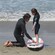 A surf instructor holding down a surfboard on the sand while a child stands on it