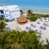 A modern 2 story building, a few thatched roof buildings and an array of recliners on a sandy beach