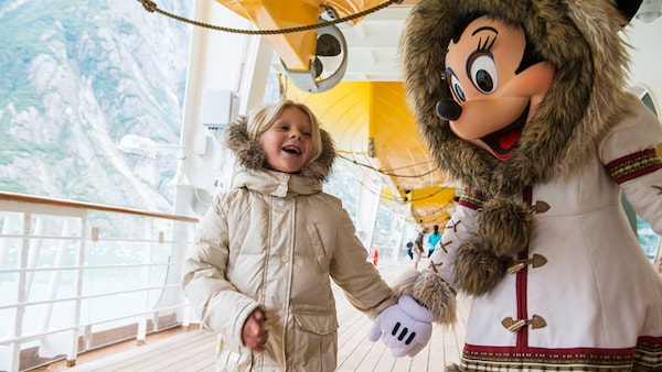 Wearing parkas, a smiling child and Minnie Mouse hold hands on the deck of a Disney Cruise Line ship