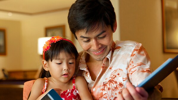 A man in a Hawaiian shirt reads a book to his young daughter