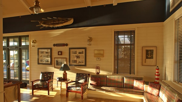 A living room with pale wood walls and floors, sofas, armchairs and a patio beyond French doors 