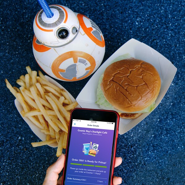 A phone showing a mobile food order confirmation from Cosmic Rays Starlight Café with a burger, fries and a drink inside a BB 8 souvenir cup