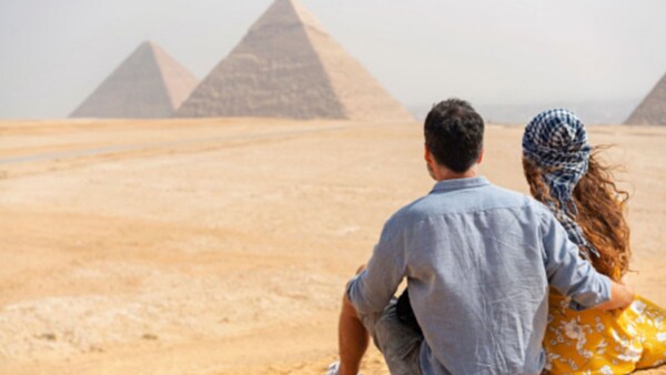 Two people sit in the sand and look at pyramids
