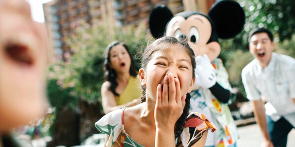 A young girl covers her mouth with her hand as she reacts in surprise with her parents and Mickey Mouse looking on behind her
