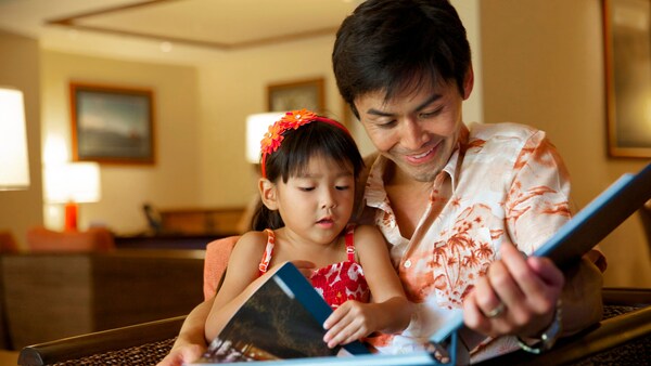 A man in a Hawaiian shirt reads a book to his young daughter