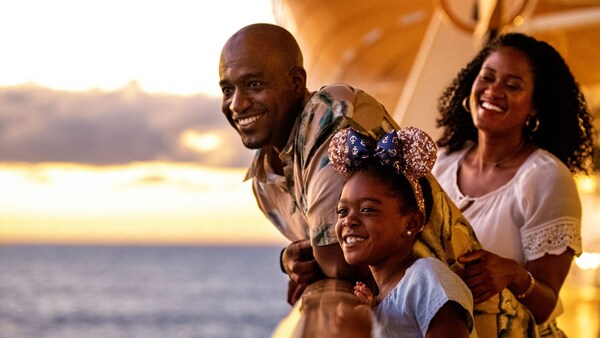 A couple and their young daughter, who wears Mickey ears, stand at the rail on the deck of a Disney Cruise Line ship, looking at the ocean waters