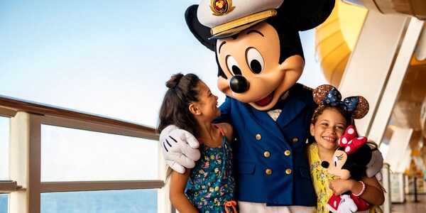 Mickey Mouse dressed as a ship's captain, hugging 2 young, smiling girls on a deck of a Disney Cruise Line ship