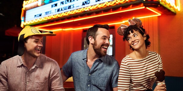 Two men, one wearing a Pluto cap, and a woman wearing Mickey ears, share a happy moment in the parks, at night