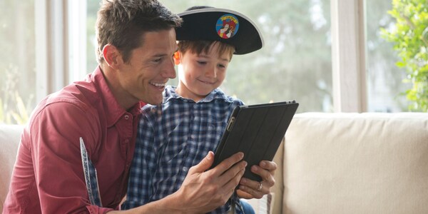Wearing a Captain Hook pirate hat, a young boy looks at a mobile device with his father 