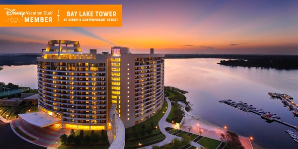 Bay Lake Tower, brightly lit at dusk, and Bay Lake, over which a banner reads, ‘Disney Vacation Club Member, Bay Lake Tower at Disney’s Contemporary Resort’