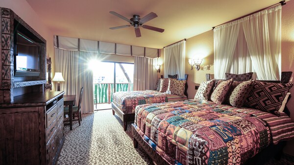 Una habitación decorada con temática africana con 2&nbsp;camas Queen Size, alfombra con estampado de animal, escritorio con una silla, centro de entretenimiento ornamentado con una cómoda de cajones, ventilador de techo y puertas de vidrio corredizas que dan a un patio al aire libre