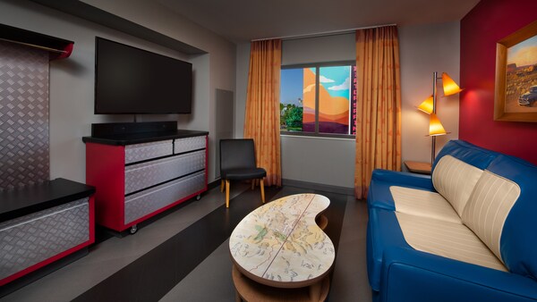 Interior de um quarto com piso laminado, uma cadeira estofada, uma mesa de centro, um sofá, uma TV de tela plana com suporte de parede, uma cômoda, um guarda-roupa, uma mesa lateral, cortinas e uma janela