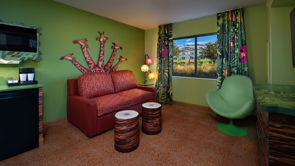 Interior de um quarto com piso laminado, uma cadeira estofada, 2 pufes, um sofá, uma TV de tela plana montada na parede, uma mesa lateral, uma cômoda, um micro-ondas, um frigobar, cortinas e uma janela